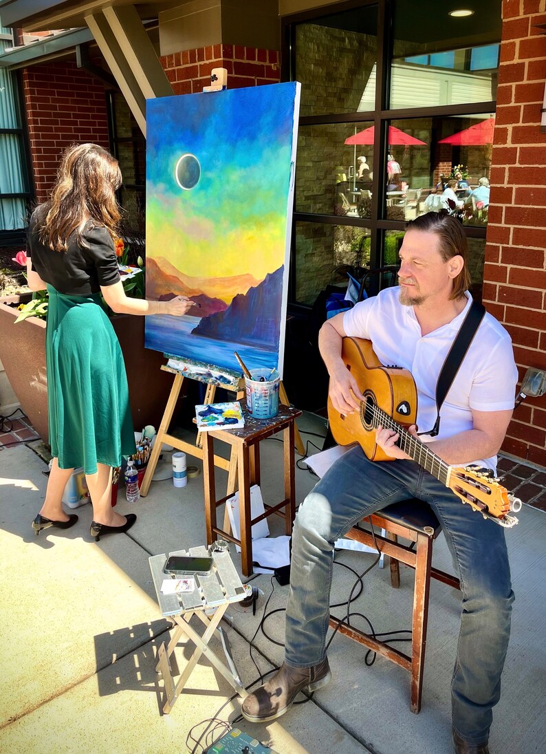 May include: A woman paints a landscape on canvas while a man plays an acoustic guitar. The painting depicts a sunset with a dark circle in the sky, possibly a solar eclipse. The scene is set outdoors, with brick walls and a window in the background.