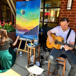 May include: A woman paints a landscape painting with a sunset and a solar eclipse in the sky. A man plays a classical guitar in the background.