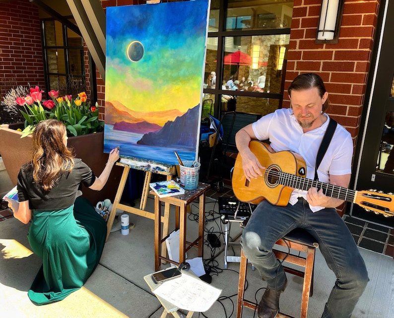May include: A woman paints a landscape painting with a sunset and a solar eclipse in the sky. A man plays a classical guitar in the background.