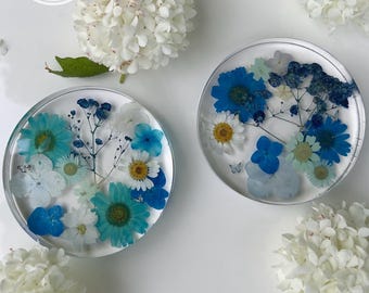 Puede incluir: Dos posavasos circulares de resina con flores secas incrustadas en tonos azules, turquesas y blancos. El arreglo floral incluye margaritas, hortensias y paniculata. Tres flores de hortensia blancas están colocadas alrededor de los posavasos.