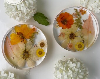 Puede incluir: Dos posavasos circulares de resina transparente que contienen flores prensadas amarillas y naranjas, follaje verde y hojas esqueletizadas. El texto SET OF TWO está impreso en un círculo blanco. Flores de hortensia blancas rodean los posavasos.