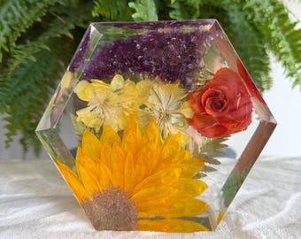 Puede incluir: Un bloque hexagonal de resina transparente que contiene flores preservadas, incluyendo un gran girasol amarillo en la base, una rosa naranja brillante, flores amarillas más pequeñas y un grupo de cristales de amatista morados incrustados.