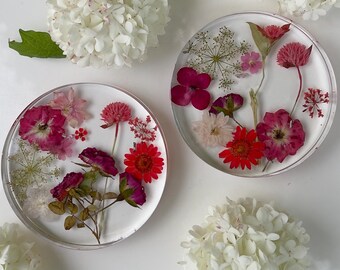 Puede incluir: Dos posavasos circulares de resina transparente que contienen flores secas prensadas en tonos rosa, rojo y blanco, dispuestos junto a tres racimos de flores de hortensia blancas frescas sobre una superficie blanca.