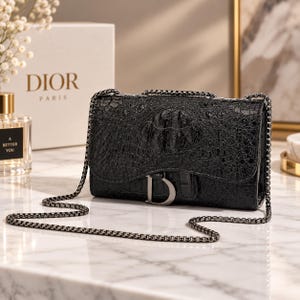 May include: A black handbag with a glossy, textured crocodile-skin pattern and a silver-toned metal chain strap. The front flap features a prominent silver-toned letter D hardware piece. The bag has a rectangular shape and a structured design.