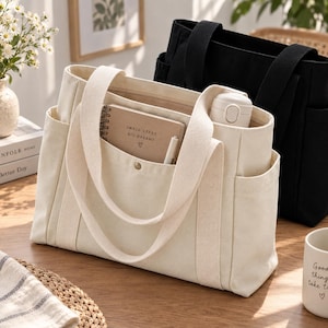 May include: A cream-coloured canvas tote bag with two sturdy handles, side pockets, and a front pocket containing a spiral-bound notebook with the text "SMALL STEPS BIG DREAMS" and a pen. A matching black canvas tote bag sits directly behind it.