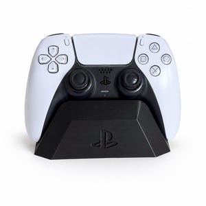 May include: A white and black wireless video game controller resting in a black charging dock. The controller features a central touchpad, two analog sticks, a directional pad, and four action buttons with triangle, circle, cross, and square symbols.