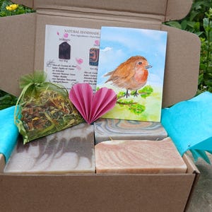 May include: A cardboard gift box containing three rectangular soap bars with marbled patterns, a green organza bag filled with dried flower petals, a pink paper heart, and a card featuring a watercolor illustration of a robin bird.