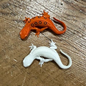 May include: Two small lizard figurines. One is bright orange with a series of black circular spots along its back and sides. The other is solid white with a smooth, unpatterned surface. Both have four legs and a curved tail, depicted in a crawling pose.