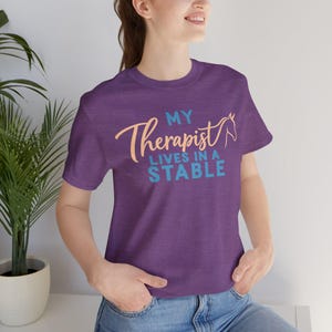 Op de afbeelding: Een paars t-shirt met korte mouwen en de tekst "MY THERAPIST LIVES IN A STABLE" in lichtblauw en perzik. Het woord "Therapist" is geschreven in een cursief lettertype en een eenvoudige lijntekening van een paardenkop is geïntegreerd.