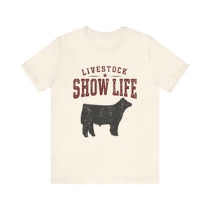 Può includere: Una t-shirt a maniche corte color crema con una grafica rosso scuro che riporta il testo "LIVESTOCK SHOW LIFE" sopra la sagoma nera di una mucca. Il design presenta una texture usurata e la maglietta ha un girocollo.