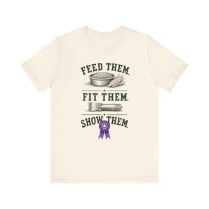 Op de afbeelding: Een crèmekleurig t-shirt met een afbeelding en de tekst FEED THEM, FIT THEM, SHOW THEM. Het ontwerp bevat illustraties van een voerbak met maïs, een tondeuse en een paars lint met een ster in het midden als prijsuitreiking.