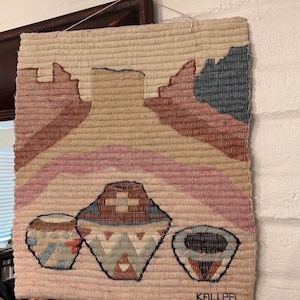May include: A rectangular woven wall hanging featuring a landscape design with a large central vase shape, layered hills in shades of pink, tan, and blue, and three smaller patterned vessels at the bottom. The text KALLPA is visible at the bottom right.
