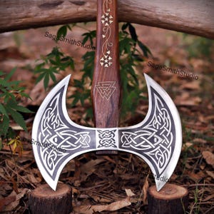 May include: A double-bitted axe with a dark wood handle featuring gold-colored floral inlays and a Valknut symbol. The metal axe head displays intricate black and silver Celtic knotwork patterns. The text SagaSmithStudio appears on the image.
