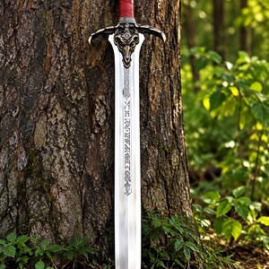 Pode incluir: Uma espada de metal prateado com punho revestido a couro vermelho, pomo em forma de coroa e guarda decorada com um motivo metálico de crânio de carneiro. A lâmina apresenta padrões rúnicos intrincados gravados ao longo do eixo central.