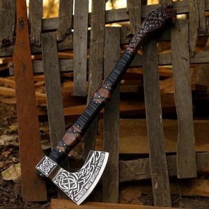 May include: A decorative axe featuring a dark brown wooden handle with black leather wrapping and a carved wolf head pommel. The metal blade displays intricate white Celtic knotwork engravings and the text "SCORCHED WOLF MENTALITY PREVAILS".