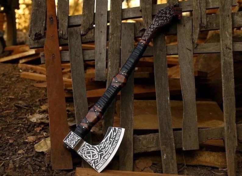 May include: A decorative axe featuring a dark brown wooden handle with black leather wrapping and a carved wolf head pommel. The metal blade displays intricate white Celtic knotwork engravings and the text "SCORCHED WOLF MENTALITY PREVAILS".
