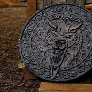 May include: A circular dark wood shield featuring a deeply carved relief of a snarling wolf head at the center. The border is decorated with intricate knotwork patterns and small metal studs spaced evenly around the outer edge of the shield.