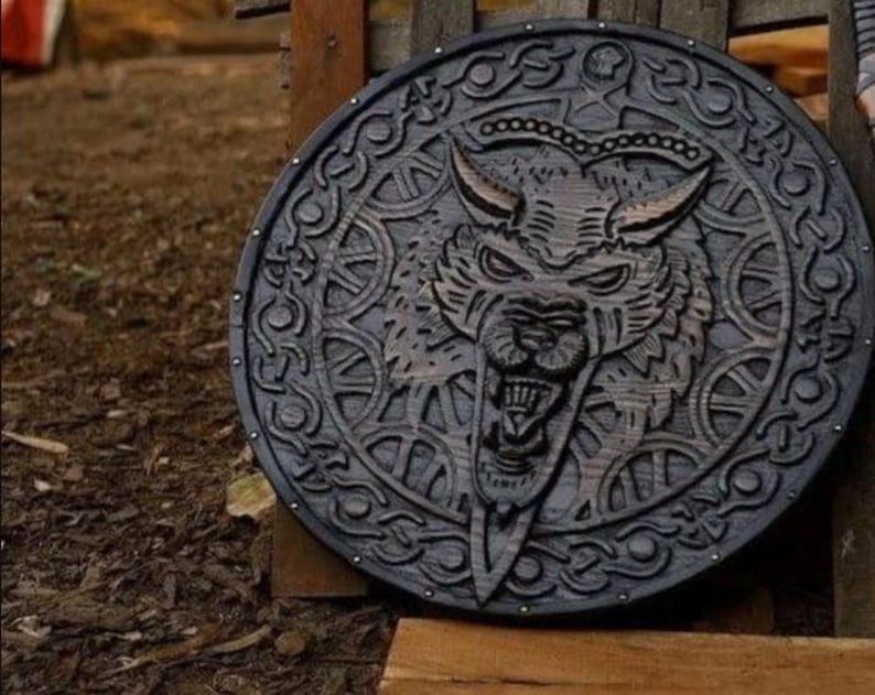 May include: A circular dark wood shield featuring a deeply carved relief of a snarling wolf head at the center. The border is decorated with intricate knotwork patterns and small metal studs spaced evenly around the outer edge of the shield.