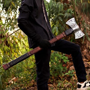 May include: A double-bit axe featuring a silver-colored head with intricate engraved knotwork patterns. The long handle is constructed from brown wood with black wrapped grip sections and metallic rivets, held horizontally across the body.