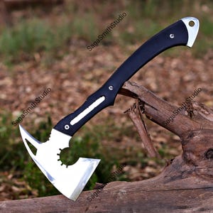 May include: A tactical hatchet with a polished silver metal head featuring a circular cutout and a sharp blade. The handle is black with silver rivets and a curved end containing a lanyard hole. The text SagaSmithStudio appears on the image.