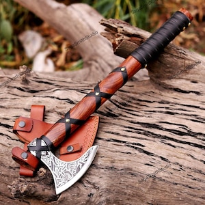 May include: A hand axe featuring a polished metal blade with intricate etched patterns, a wooden handle wrapped in black leather strips secured with brass rivets, and a matching tan leather sheath with snap closures and visible stitching.