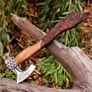 May include: A hand-axe with a dark brown wooden handle featuring intricate carved patterns and a wolf-head motif. The metal axe head displays a silver finish with detailed engravings of a wolf and decorative knotwork on the blade surface.