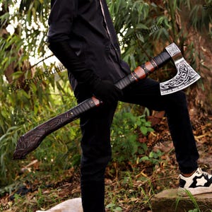 May include: A long-handled battle axe featuring a dark wooden shaft with intricate carved patterns and black leather wrapping. The metal axe head displays an engraved Celtic knot design and the text "SCORCHED WOLF MENTALITY PREVAILS".