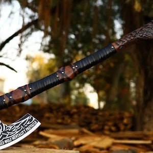 May include: A metal axe with a silver blade featuring intricate white Celtic knotwork and the text "SCORCHED WOLF MENTALITY PREVAILS". The handle is made of dark wood wrapped in black leather cord, ending in a carved wooden wolf head pommel.
