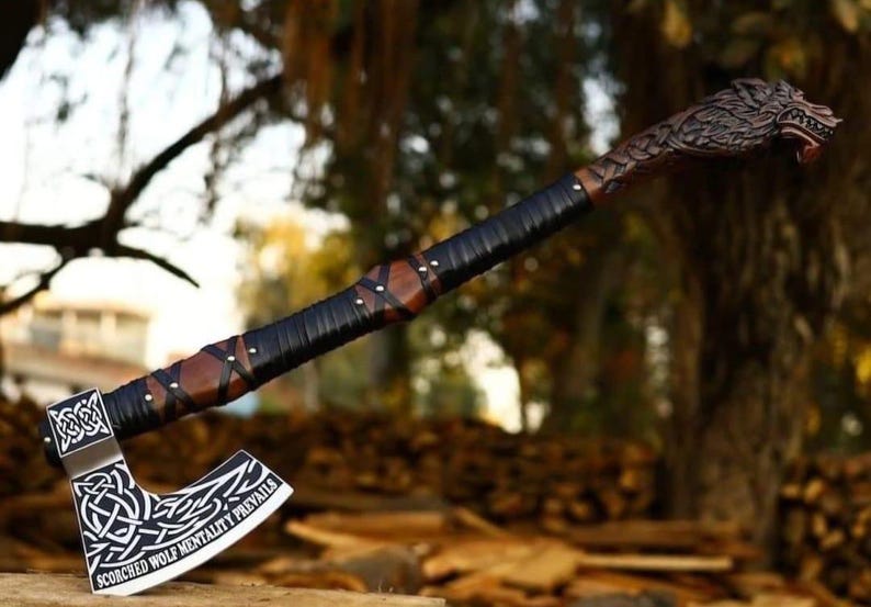 May include: A metal axe with a silver blade featuring intricate white Celtic knotwork and the text "SCORCHED WOLF MENTALITY PREVAILS". The handle is made of dark wood wrapped in black leather cord, ending in a carved wooden wolf head pommel.