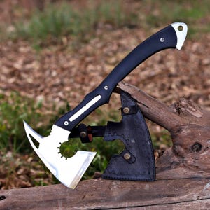 Può includere: Un'ascia tattica con una testa in metallo argentato lucido caratterizzata da un design traforato e un manico nero curvo. L'ascia è parzialmente inserita in un fodero in pelle nera con cuciture decorative e due bottoni a pressione in metallo ottonato.