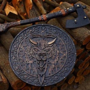 May include: A circular wooden shield featuring a carved wolf head at the center surrounded by intricate knotwork patterns, paired with a long-handled axe that has a dark leather sheath covering the blade and a carved wolf head on the handle.