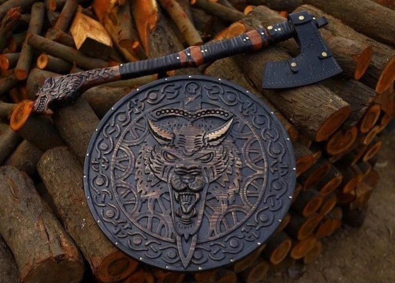 May include: A circular wooden shield featuring a carved wolf head at the center surrounded by intricate knotwork patterns, paired with a long-handled axe that has a dark leather sheath covering the blade and a carved wolf head on the handle.
