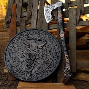 May include: A circular black wooden shield featuring a carved wolf head design with intricate knotwork patterns, paired with a long-handled axe. The axe has a silver metal head with engraved knotwork and a dark leather-wrapped handle with a carved pommel.
