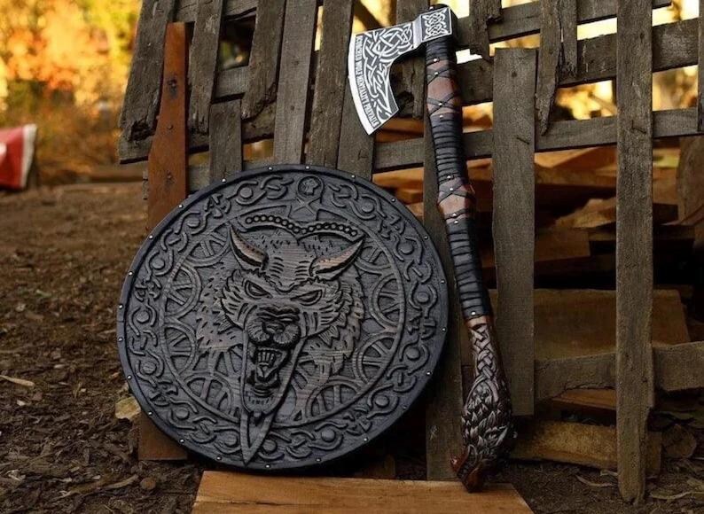 May include: A circular black wooden shield featuring a carved wolf head design with intricate knotwork patterns, paired with a long-handled axe. The axe has a silver metal head with engraved knotwork and a dark leather-wrapped handle with a carved pommel.