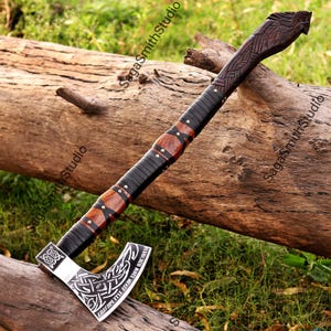 Può includere: Un'ascia a mano con una lama in metallo lucido con intricati intagli a nodo celtico e il testo "VICTORY OR VALHALLA, MIGHTY PREVAILS". Il manico è realizzato in legno scuro con rivestimento in pelle nera, rivetti metallici e un pomolo intagliato.
