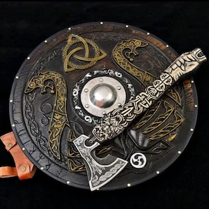 Op de afbeelding: Een rond donkerbruin houten schild met goudkleurige reliëfsnijwerken van slangen en een triquetra, met een centrale zilverkleurige metalen naaf. Een zilverkleurige metalen bijl met ingewikkelde gegraveerde patronen ligt diagonaal over het schild.