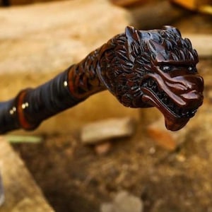 May include: A hand axe featuring a dark brown wooden handle carved into the shape of a wolf head at the pommel. The handle is wrapped in black leather cord with small brass rivets, leading to a metal axe head with an etched blade surface.