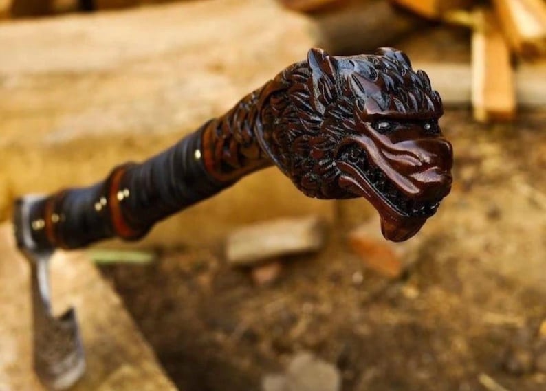 May include: A hand axe featuring a dark brown wooden handle carved into the shape of a wolf head at the pommel. The handle is wrapped in black leather cord with small brass rivets, leading to a metal axe head with an etched blade surface.