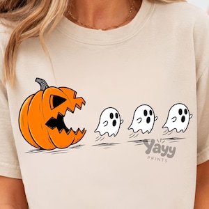 May include: A beige crewneck sweatshirt featuring a graphic print of a large orange jack-o'-lantern with a jagged mouth chasing three small white ghosts. The text "Yayy PRINTS" is printed in small grey letters below the ghosts on the shirt.