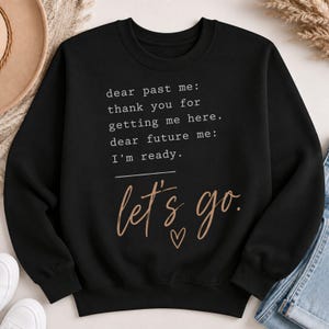 Może przedstawiać: Czarna bluza z długim rękawem z białym napisem: dear past me: thank you for getting me here. dear future me: I'm ready. Poniżej słowa let's go. są nadrukowane beżową czcionką imitującą pismo odręczne z małą ikoną serca.