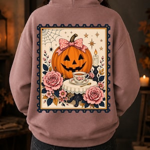 May include: A mauve hoodie featuring a rectangular graphic on the back. The design shows an orange jack-o'-lantern with a pink polka-dot bow, a teacup on a lace-covered table, pink roses, a spider web, and a lit candle, all framed by a dark border.