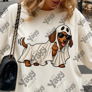 May include: A cream-colored sweatshirt featuring a graphic of a brown dachshund wearing a white ghost costume with black sunglasses. The dog is smiling with its tongue out and holding up its middle finger. The text Yayy Prints is repeated.