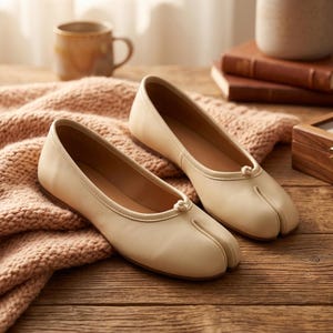 May include: A pair of cream-colored split-toe ballet flats featuring a smooth leather-like finish, a low-profile silhouette, and a small decorative knot detail at the center of the vamp. The shoes have a flat sole and a distinct cleft toe design.