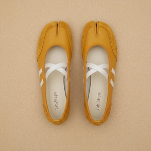 May include: A pair of mustard yellow Tabi-style split-toe flats featuring a smooth leather-like finish with suede side panels. Each shoe includes a white elastic cross-strap across the top and the brand name TabiSteps printed on the inner sole.