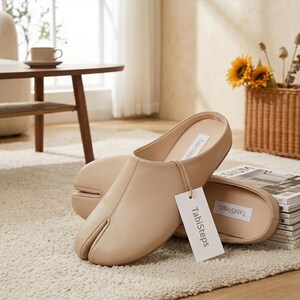 May include: A pair of beige slip-on mules with a split-toe design. The interior features a white label with the text Tabisteps. A white rectangular hang tag attached with a string also displays the text Tabisteps in a dark gray font.