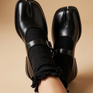 Könnte beinhalten: Ein Paar schwarze Mary-Jane-Schuhe aus Leder mit markantem Zehenschlitz-Design, einem einzelnen Riemen mit Metallschnallenverschluss und einem niedrigen Absatz. Die Schuhe werden mit schwarzen Rippsocken mit zarter Spitzenborte getragen.