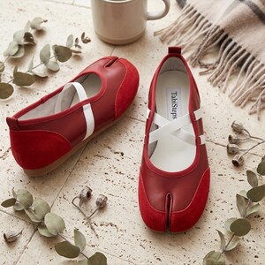 May include: A pair of red split-toe flats featuring a smooth leather and suede construction with white elastic cross-straps. The interior insole is labeled with the text TabiSteps. The shoes have a low-profile design with a distinct toe separation.