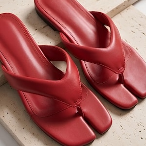 Könnte beinhalten: Ein Paar rote flache Sandalen mit einem breiten, gepolsterten Zehensteg und einem markanten Zehentrenner-Design. Die Sandalen haben eine glatte, einfarbig rote Oberfläche mit einer quadratischen Zehenpartie und einer flachen Sohle.