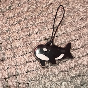 May include: A black and white orca whale charm made of a smooth, glossy material. The charm features the iconic markings of an orca, including a white eye patch and belly, and is attached to a thin, black cord loop for hanging or attachment.