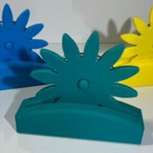 May include: Three flower-shaped card holders in blue, teal, and yellow. Each holder features a central circular detail and a wide, curved base with a horizontal slot designed to securely hold a card or paper in an upright position.